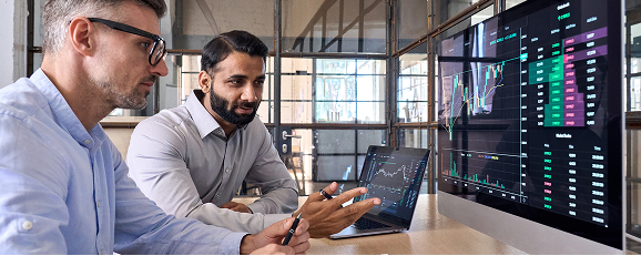 two men sitting at a table and having a discussion while looking at a computer screen displaying an analytics dashboard