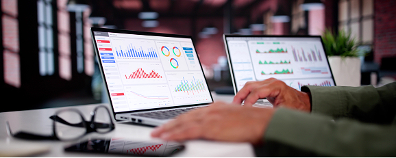 a business professional sitting at a desk and looking at analytics dashboards displayed across two laptop screens